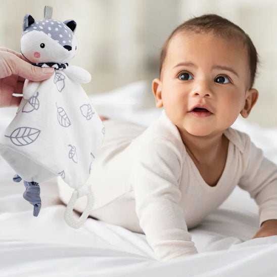 Un bébé en position ventrale, regardant une personne lui tendre un doudou peluche en forme de renard blanc avec des motifs feuilles et noisettes