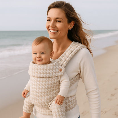 Mère souriante sur une plage portant son bébé dans un porte bébé physiologique imprimer de couleur beige et blanc