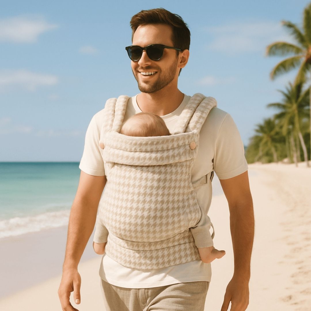 Un père marchant sur la plage en portant son enfant face contre lui dans un porte bébé physiolgoique à motif beige et blanc
