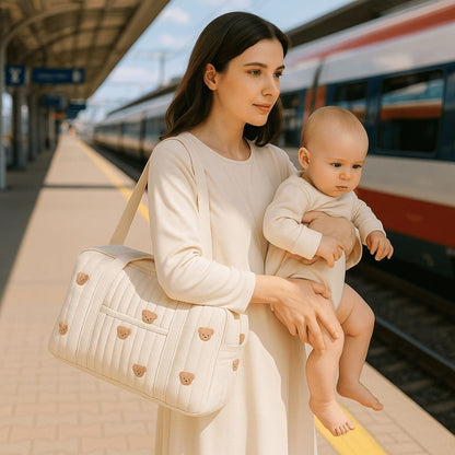 Une mère portant son bébé dans les bras sur le quai d'une gare, sac à langer beige avec des motifs de petits ours.